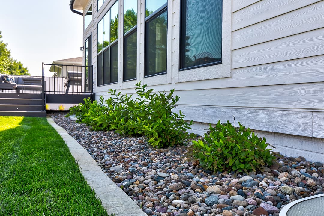 Lush green plants along a gravel border next to a modern home exterior and patio area.