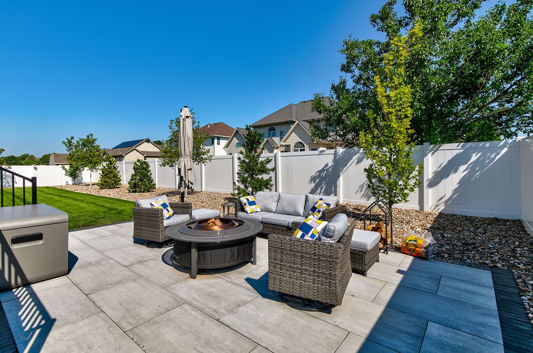 Modern backyard patio with fire pit, furniture, and lush landscaping under a clear blue sky.
