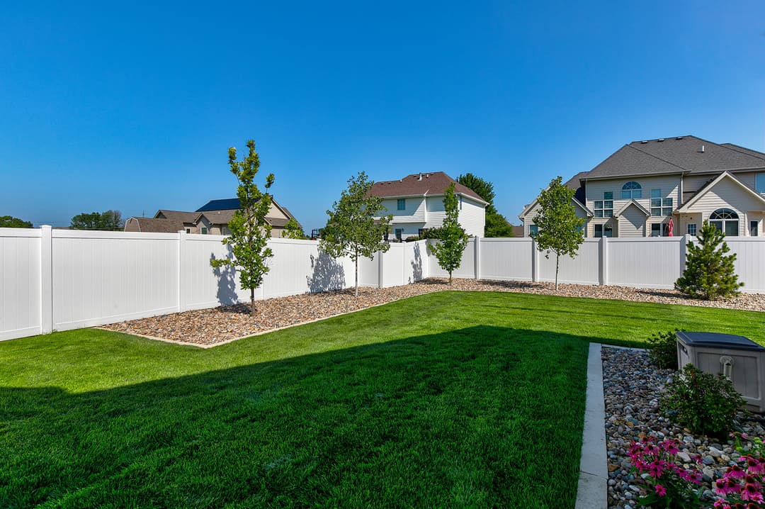 Lush green backyard with decorative trees and white vinyl fence under clear blue sky.
