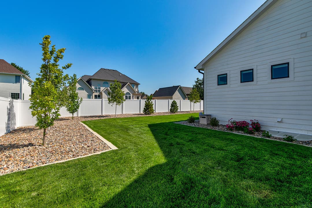 Lush green backyard with trees, stone landscaping, and white vinyl fence under clear blue sky.