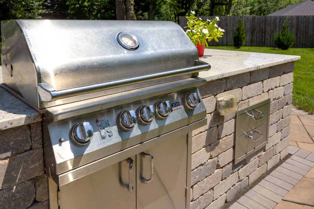 Stainless steel outdoor grill on stone patio with green lawn and potted plant nearby.