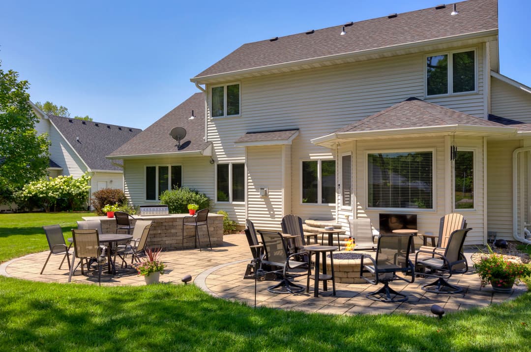 Modern home exterior with landscaped patio, seating area, and greenery in sunny backyard.