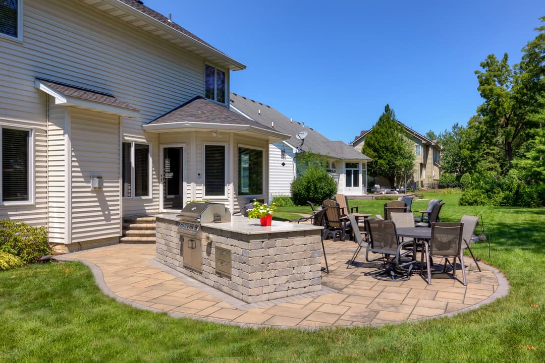 Outdoor patio with a stone barbecue, dining table, and lush green lawn in a sunny backyard.