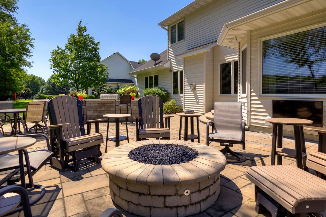 Outdoor patio with seating and a fire pit, surrounded by green trees and sunshine.