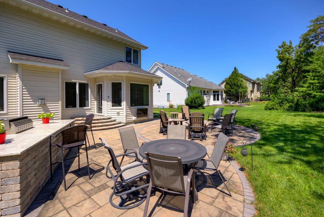 Spacious backyard patio with tables, chairs, and greenery under a clear blue sky.
