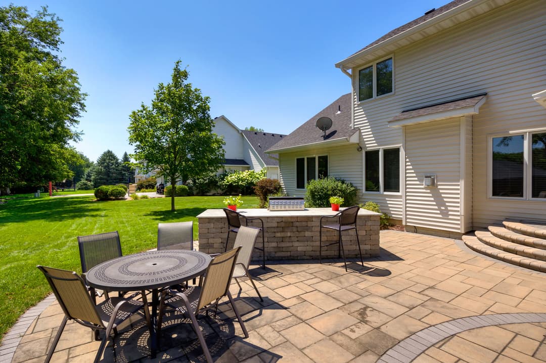 Patio dining area with stone wall, chairs, trees, and sunny backyard in suburban home setting.