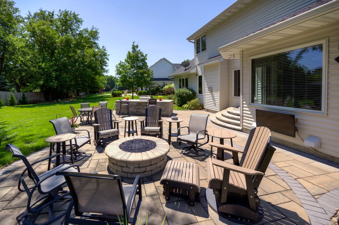 Outdoor patio with fire pit, seating, and green lawn in a sunny residential backyard.