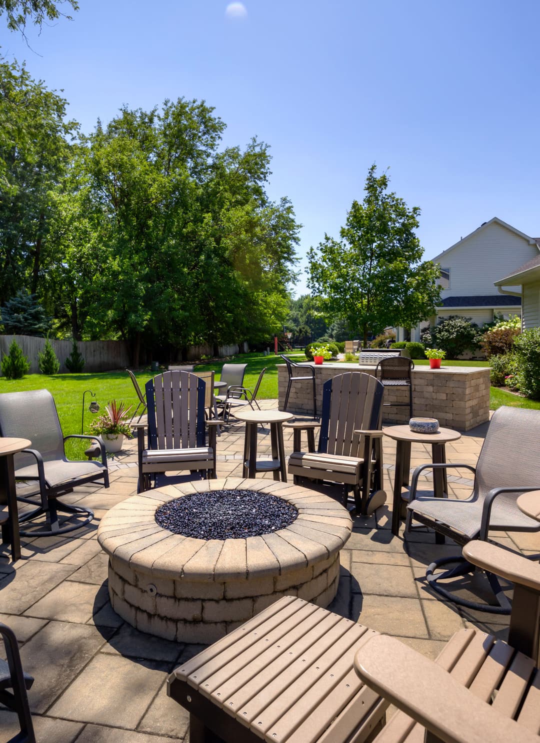 Backyard patio with seating, stone fire pit, and lush greenery under clear blue sky.