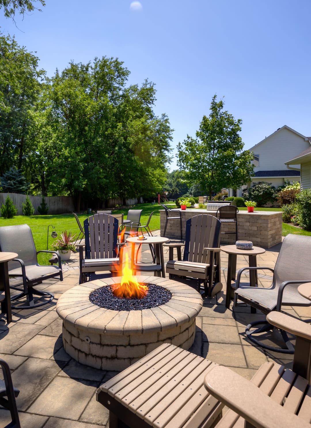 Cozy outdoor patio with a fire pit, seating area, and lush greenery under a clear blue sky.