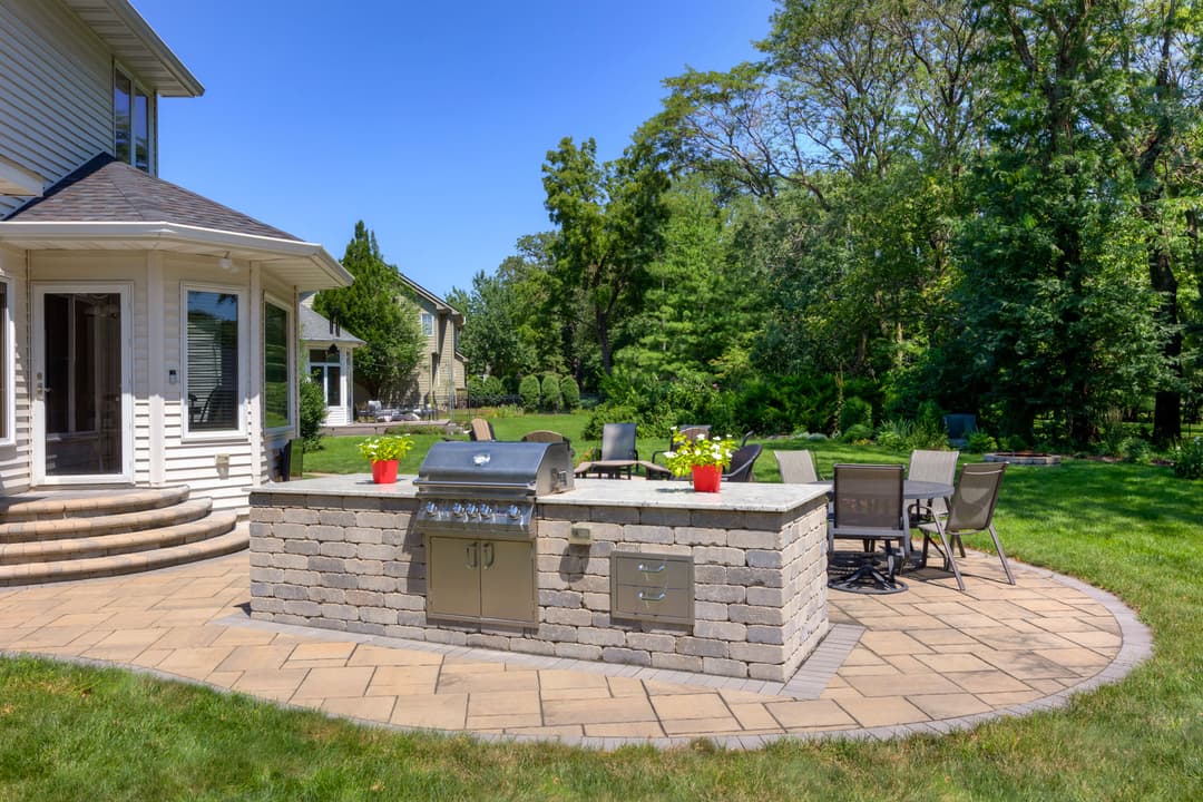 Modern outdoor kitchen with grill, patio seating, and lush greenery in a sunny backyard.