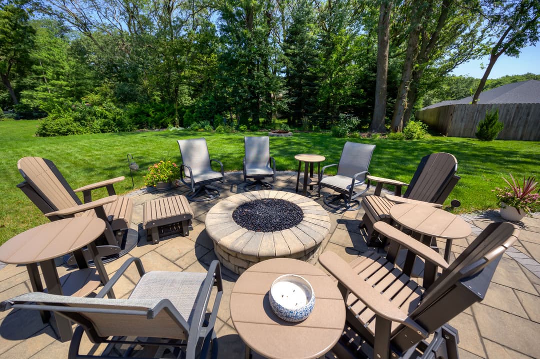Outdoor seating area with fire pit surrounded by chairs and tables in a lush garden setting.