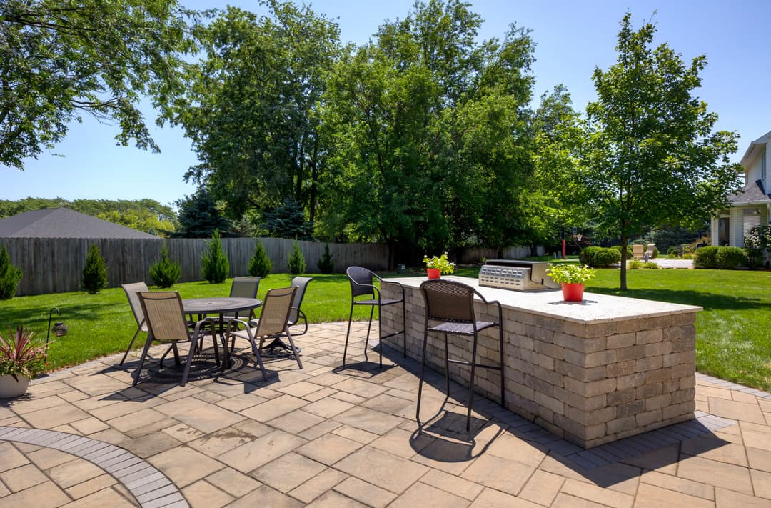 Outdoor patio featuring a stone bar, dining set, and lush green landscaping in bright sunlight.