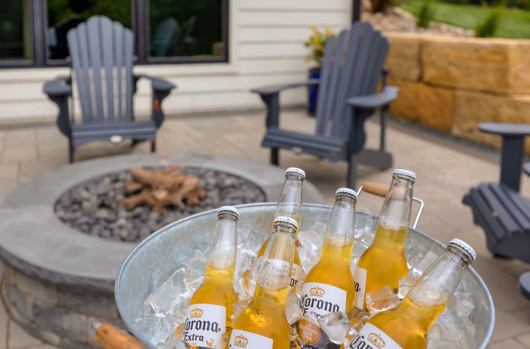 Chilled Corona beers in a bucket beside a fire pit and Adirondack chairs in a backyard setting.