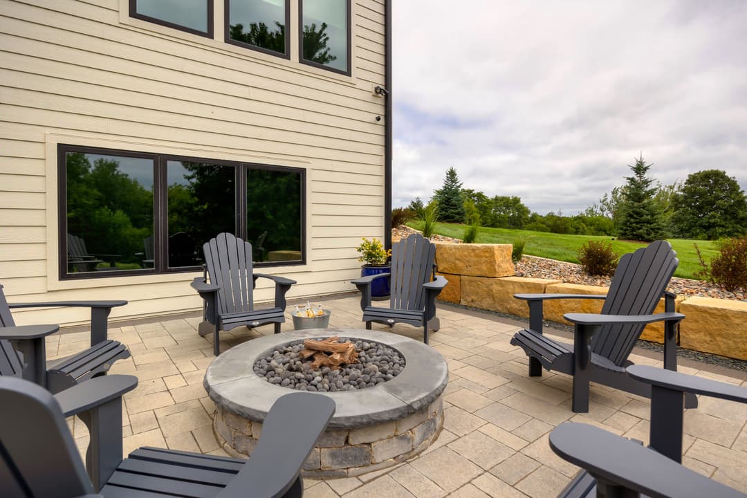 Outdoor patio with Adirondack chairs around a stone fire pit, surrounded by greenery.
