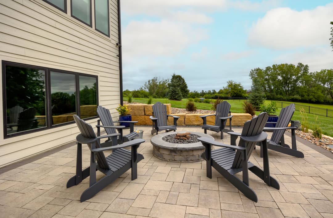 Outdoor patio with black Adirondack chairs around a stone fire pit, surrounded by greenery.