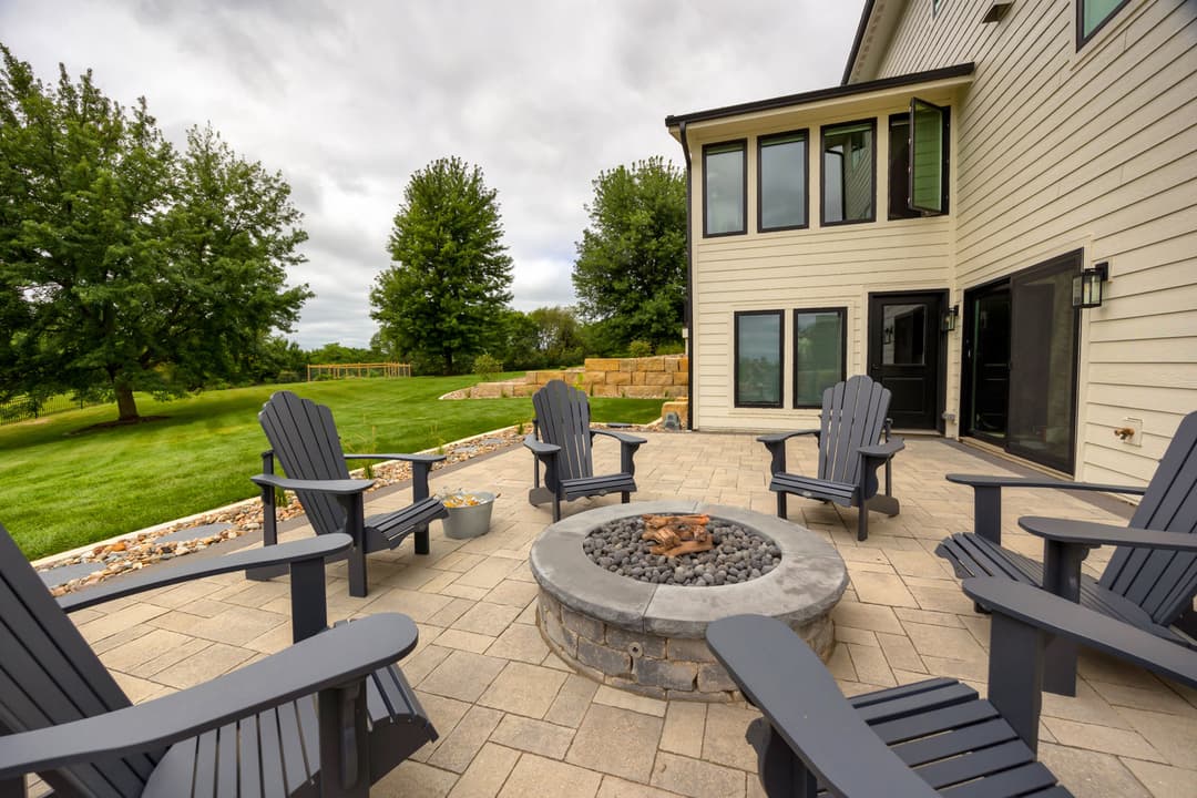 Outdoor patio featuring a stone fire pit surrounded by black Adirondack chairs and grassy landscape.