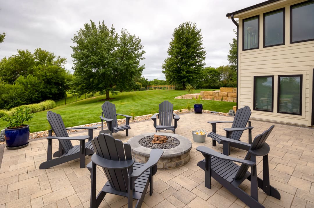 Patio featuring gray Adirondack chairs around a stone fire pit, surrounded by lush greenery.