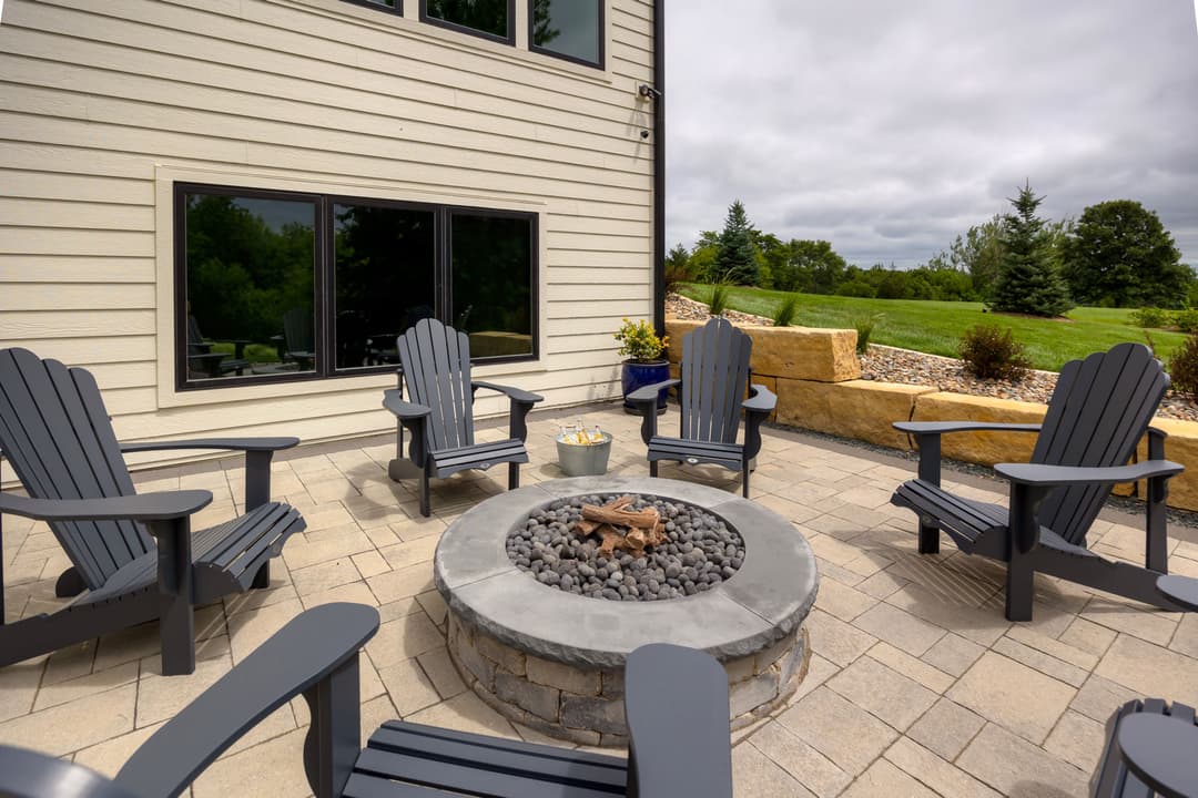 Outdoor patio with gray Adirondack chairs around a stone fire pit and green landscape backdrop.