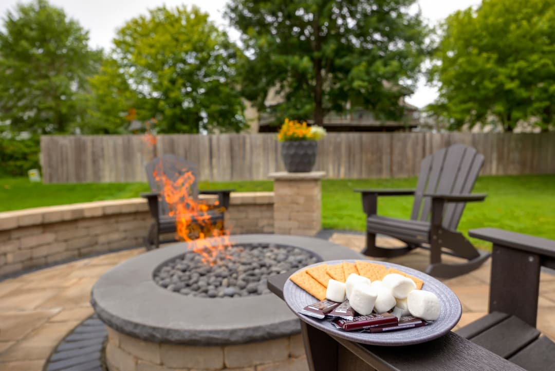 S'mores ingredients on a plate by a cozy fire pit in a backyard setting.