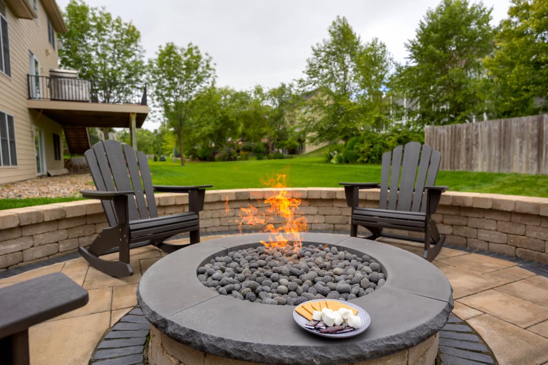 Fire pit with flames, surrounded by stones, chairs, and a plate of s'mores in a backyard.