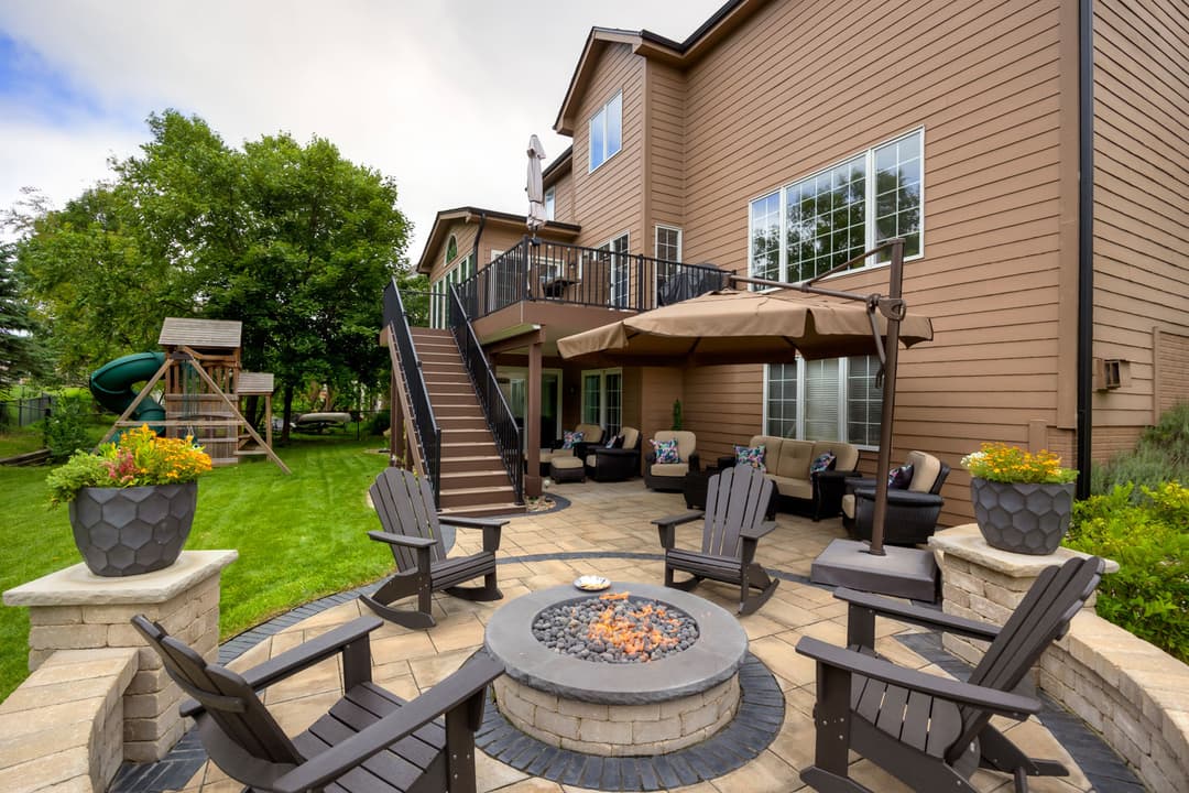 Modern backyard patio with fire pit, seating, and playset surrounded by lush greenery.