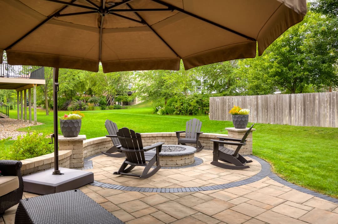 Cozy backyard patio with fire pit, Adirondack chairs, and lush green lawn under large umbrella.