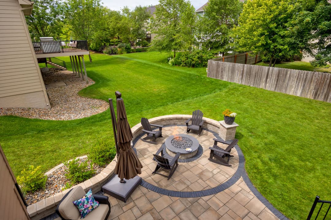 Backyard with stone fire pit, seating area, green lawn, and trees in a suburban setting.