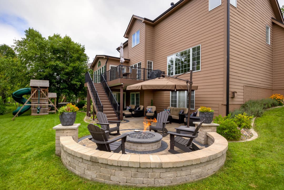 Backyard patio with fire pit, seating area, and playset, surrounded by lush greenery.