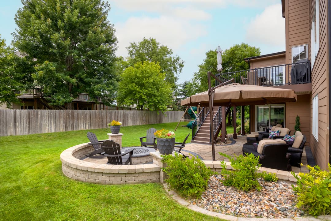 Backyard patio with seating, fire pit, and green lawn surrounded by trees and wooden fence.