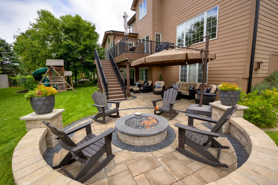 Outdoor fire pit area with seating, surrounded by landscaping and a playset in the background.