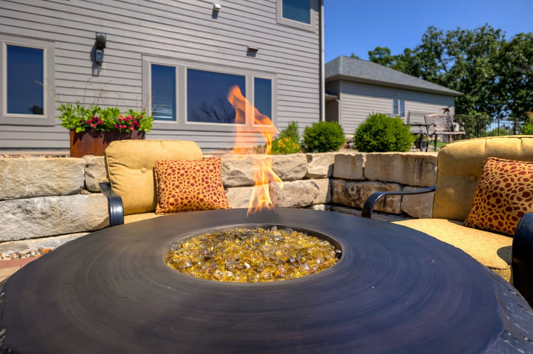 Fire pit with flame and decorative stones, surrounded by outdoor seating and a garden backdrop.