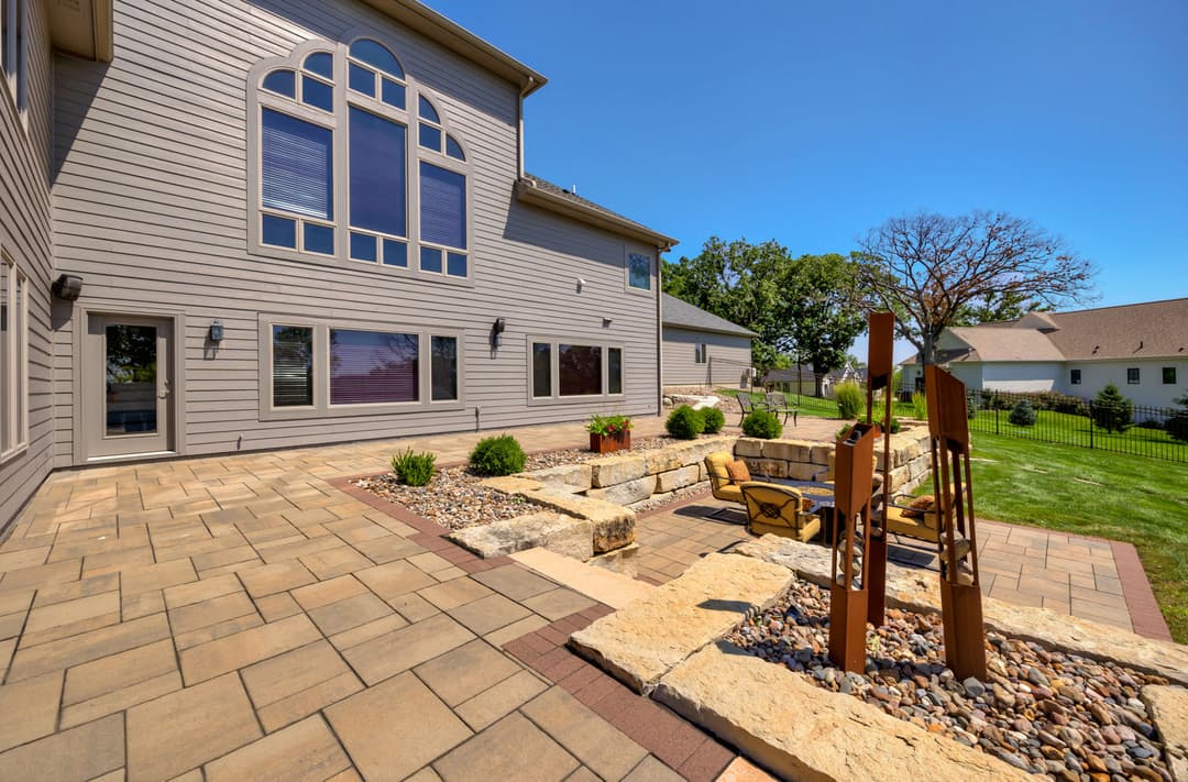 Modern backyard patio with seating, stone features, and landscaped greenery under blue skies.