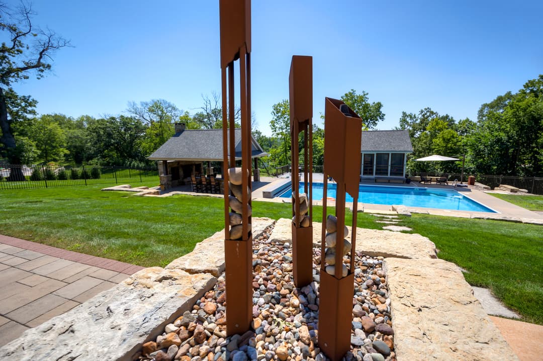 Modern outdoor sculpture surrounded by pebbles, with a pool and greenery in the background.