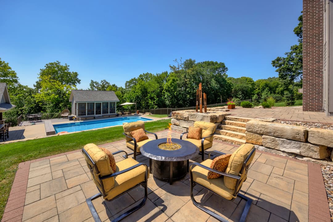 Outdoor patio with seating around a fire pit, pool, and lush landscaping in the background.