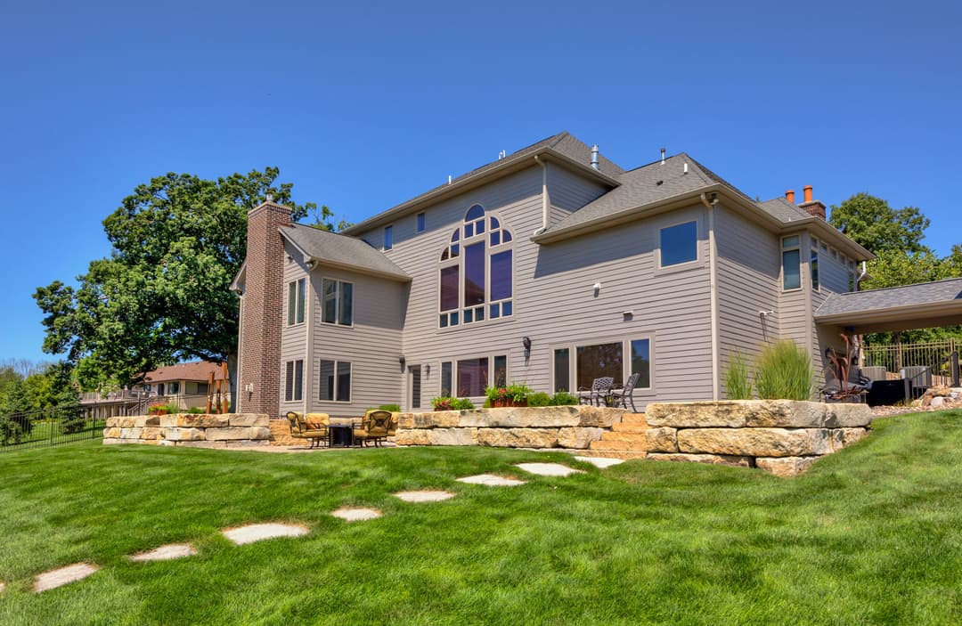 Large modern house with stone patio, lush green lawn, and clear blue sky. Ideal outdoor space.