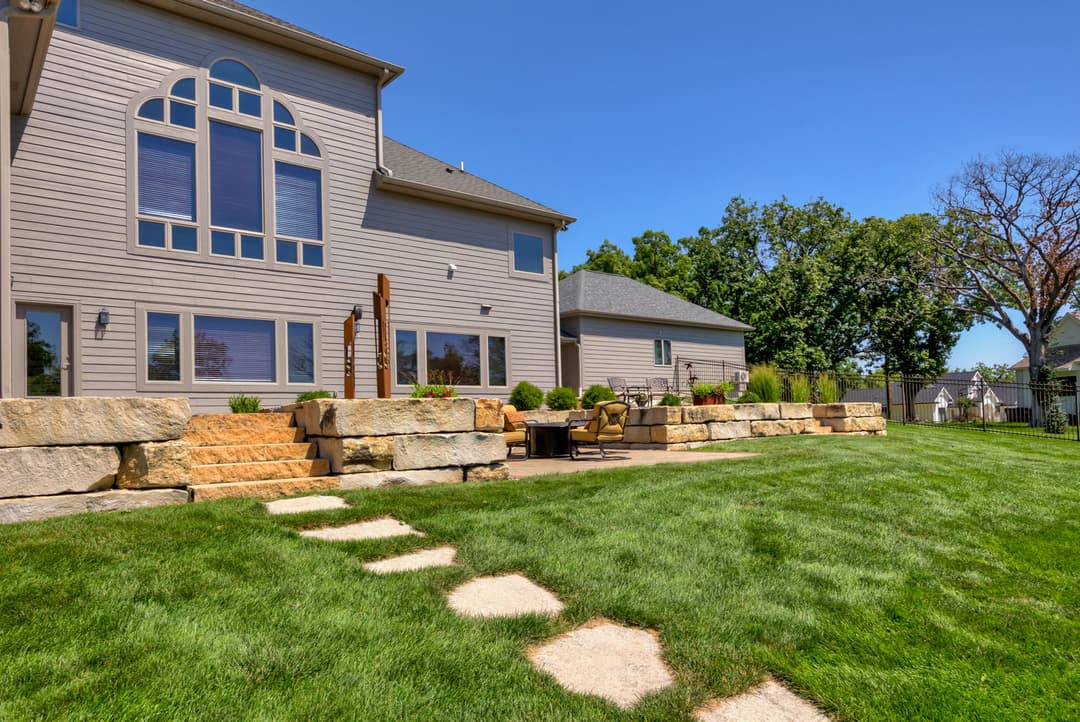 Modern home exterior with landscaped backyard, stone steps, and seating area on a sunny day.