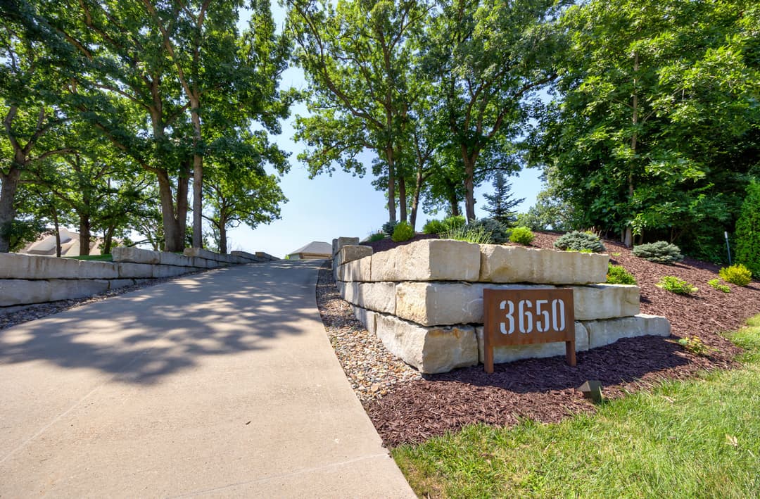 Driveway entrance with stone landscaping and house number 3650 surrounded by trees.