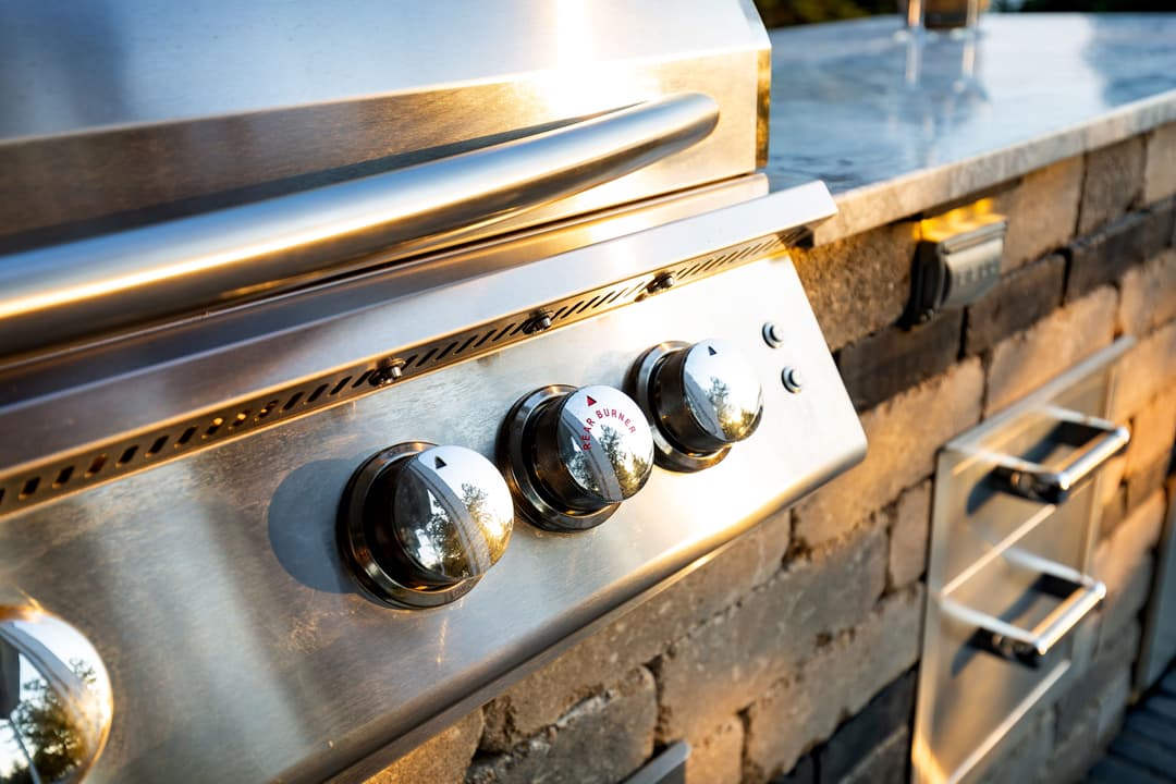 Stainless steel outdoor grill with knobs and stone countertop at sunset.