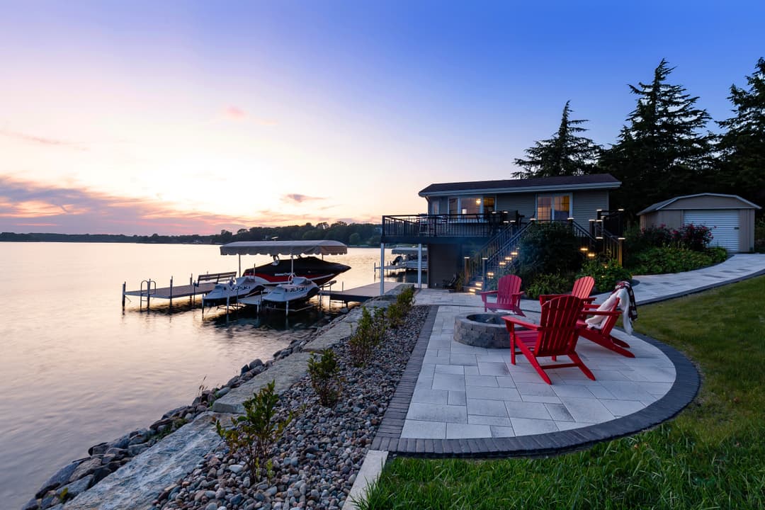 Scenic lakefront home with red chairs, docked boats, and vibrant sunset view.