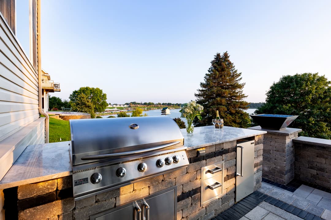 Outdoor kitchen with stainless steel grill overlooking serene lake and lush greenery.