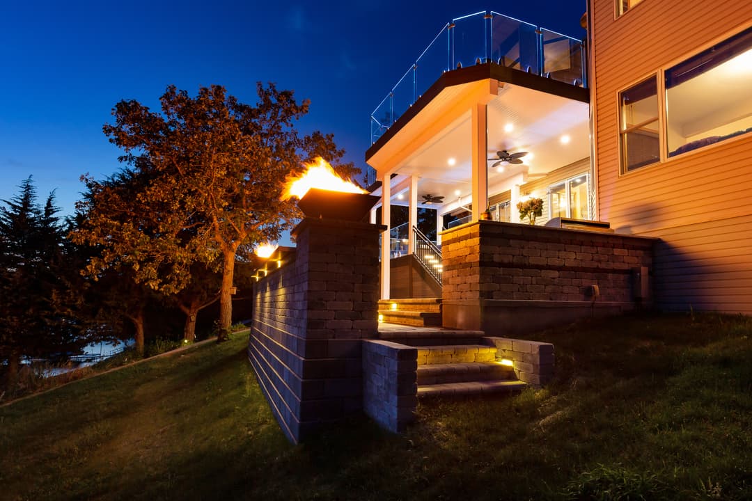 Modern home exterior with patio lighting and fire feature at dusk, surrounded by trees.