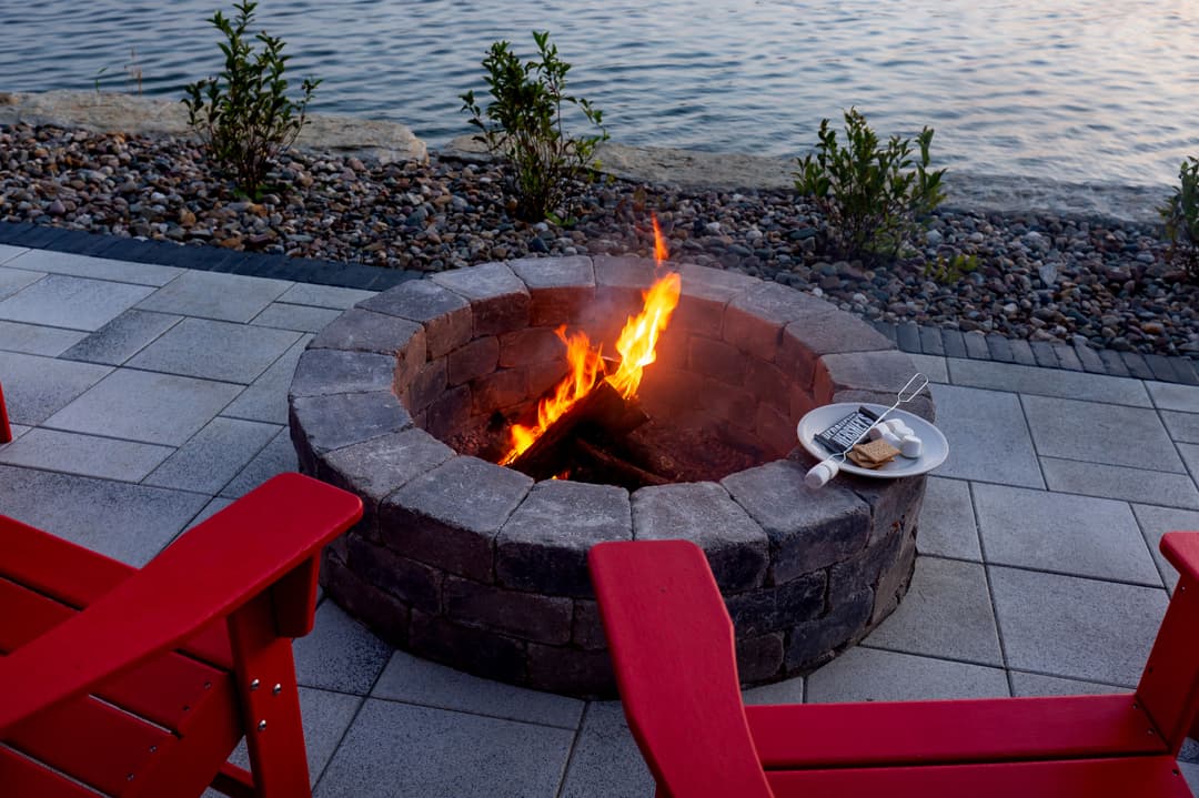 Cozy fire pit with flames, red chairs, and s'mores plate by the waterfront at sunset.