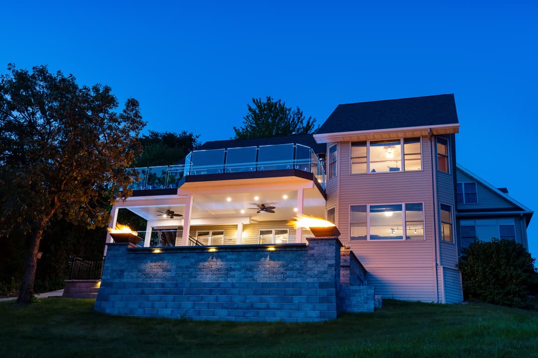 Modern two-story house with illuminated deck and patio at dusk, surrounded by greenery.