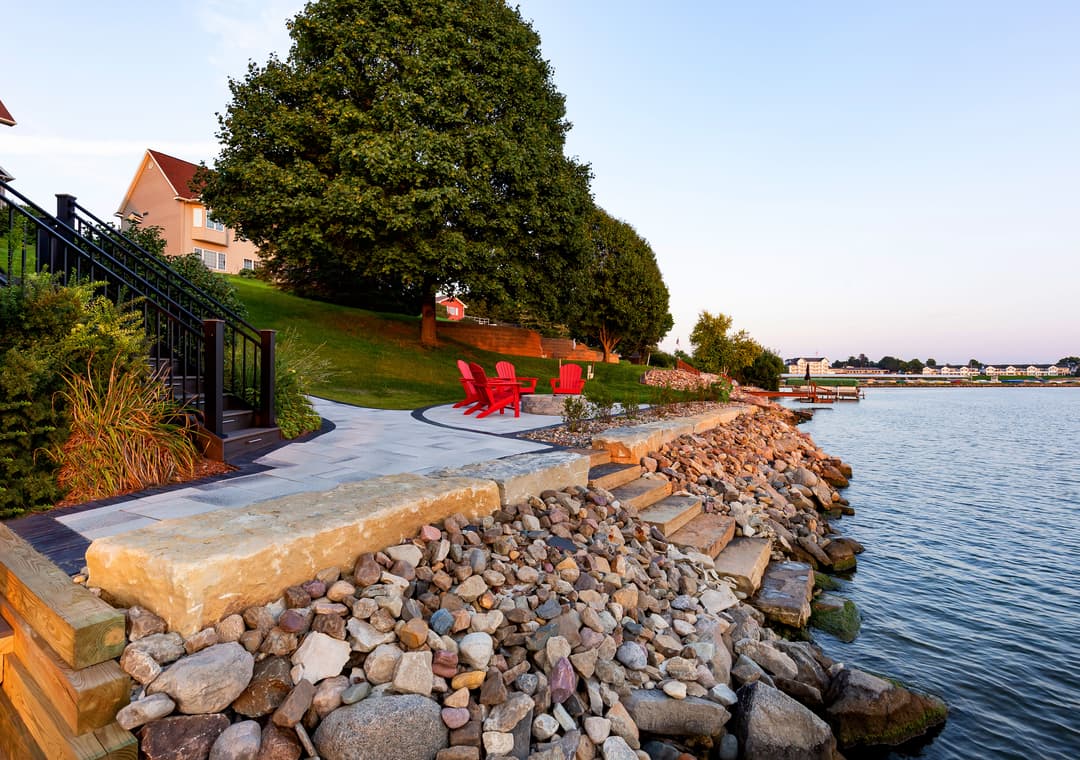 Red adirondack chairs by water's edge with stone shoreline and lush greenery at sunset.