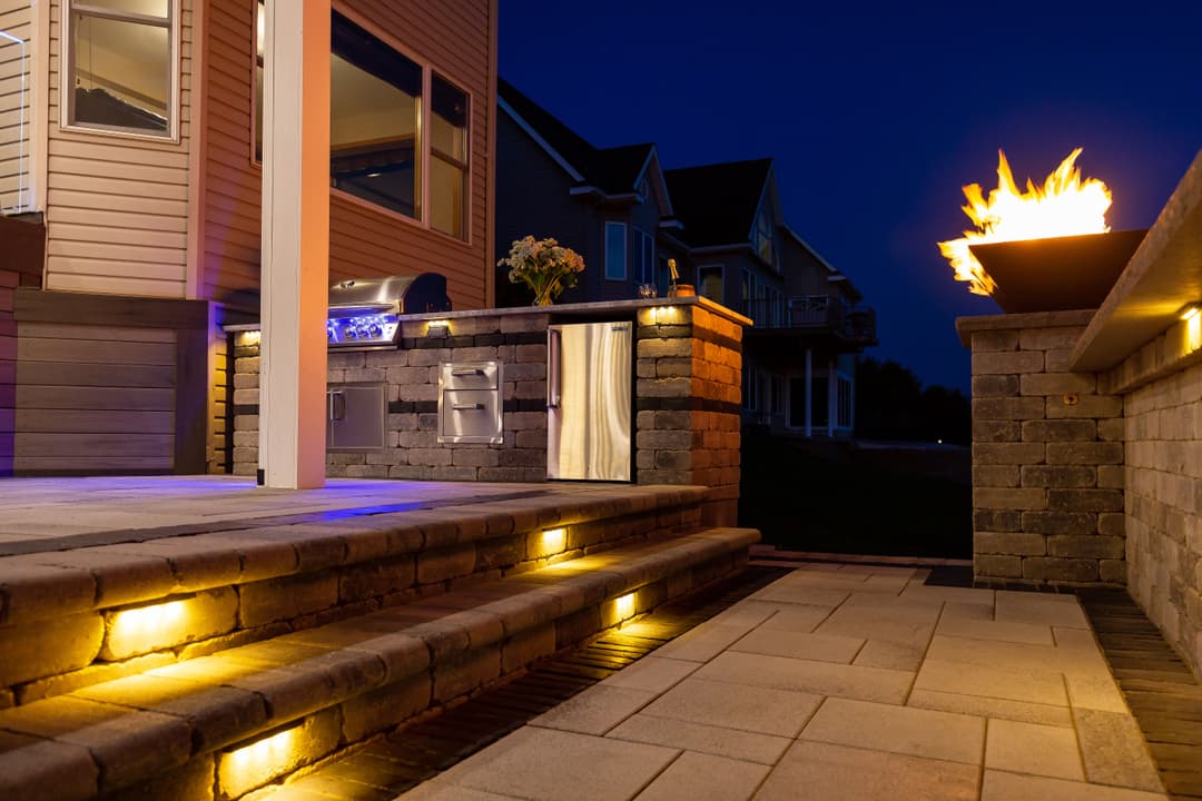 Outdoor patio at night featuring a stone grill, fire pit, and ambient lighting.