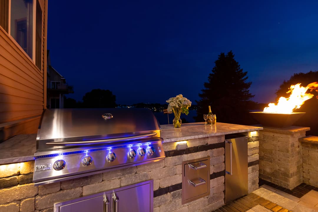 Modern outdoor kitchen with stainless steel grill, stone counter, and fire pit at dusk.