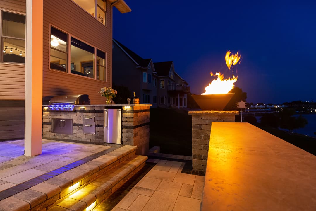 Modern outdoor kitchen with fire feature at dusk by a lakeside home. Bright lights and sunset glow.