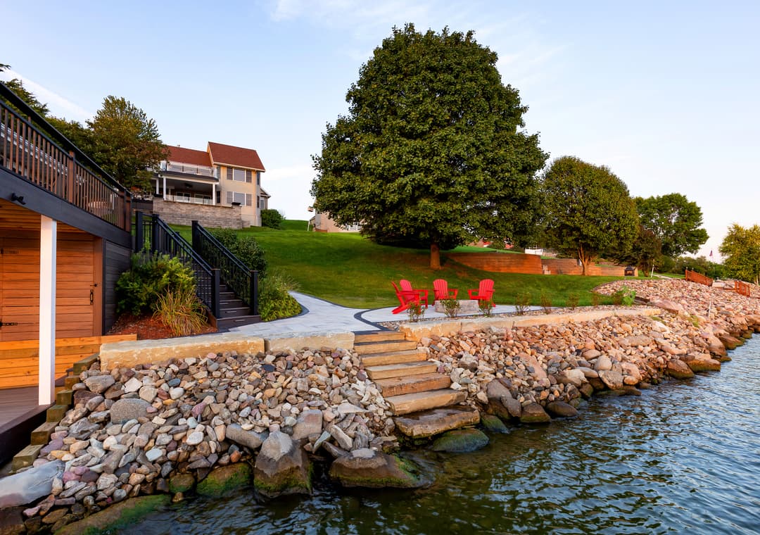 Lakeside view featuring a patio with red chairs, a stone path, and a lush green lawn.