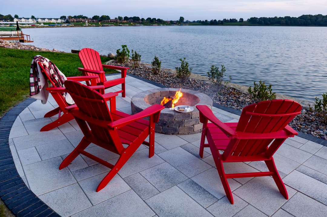 Red Adirondack chairs around a fire pit by the lake at sunset, creating a cozy outdoor setting.