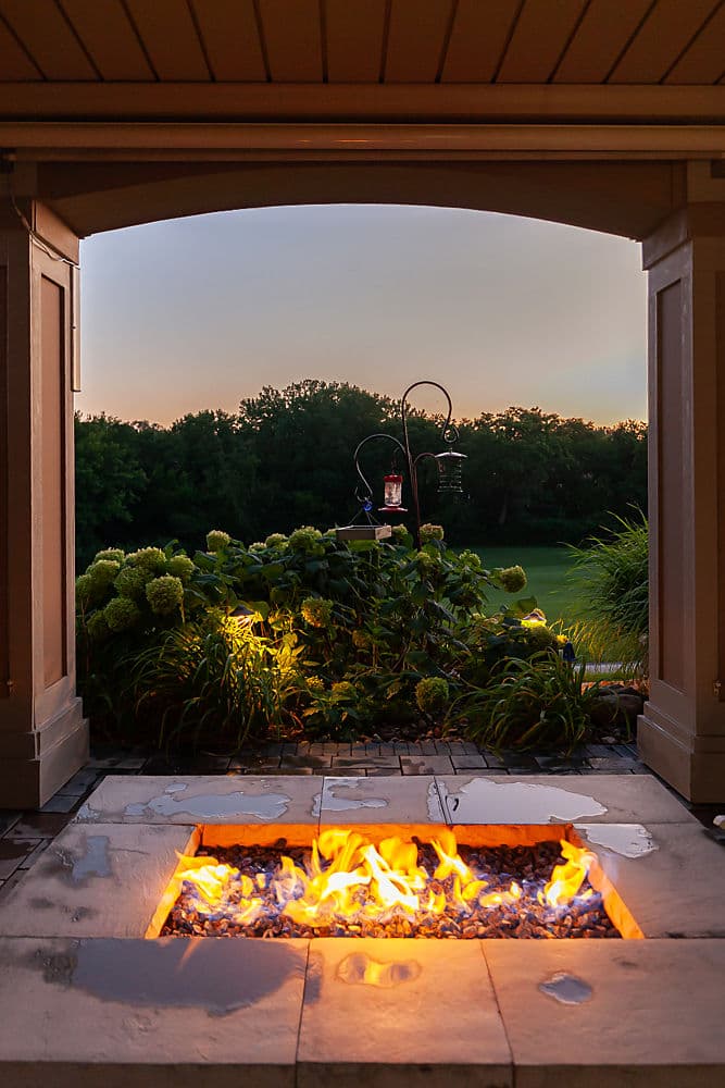 Outdoor fire pit glowing at dusk with lush greenery and scenic landscape view.